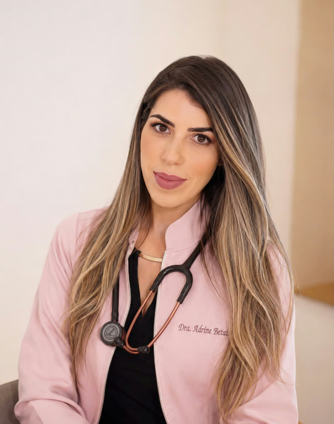 Dra. Adrine Betat, de jaleco rosa e estetoscópio, olhando para a câmera